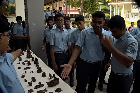 Students gathered around a long table of small clay figures, one pointing