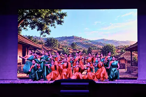 A large group of dancers in red, green and teal costumes posed together on stage, in front of a screen showing a village below green hills