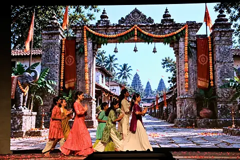 Dancers in red, cream and green costumes on stage, in front of a screen showing a carved stone gateway hung with marigold garlands