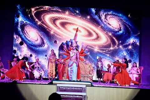 Performers in red and orange costumes on stage, in front of a screen showing a spiral galaxy, with a many-armed figure at the centre