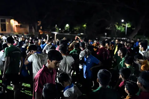 Students crowded on a lawn at night under the trees and lamps