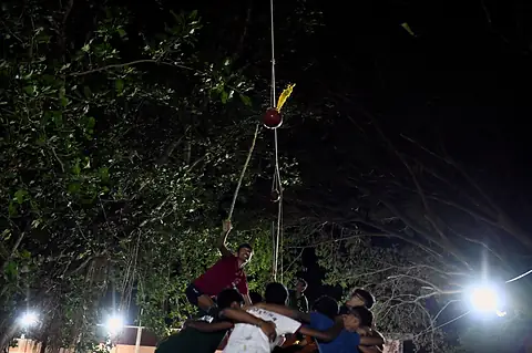 Students building a human pyramid under a tree at night, a pot hanging above them
