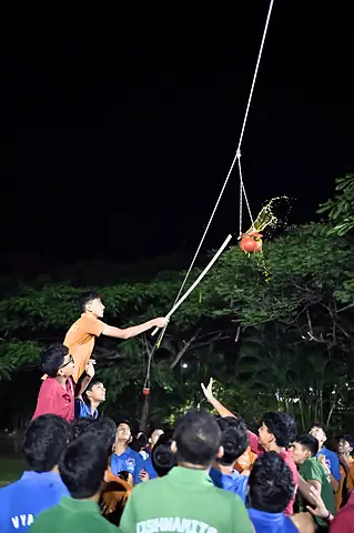 A student on the shoulders of others reaching up for a clay pot hung from a rope, after dark