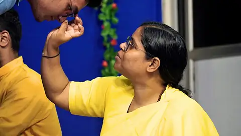 A woman in a yellow sari touching another person’s forehead before a blue backdrop
