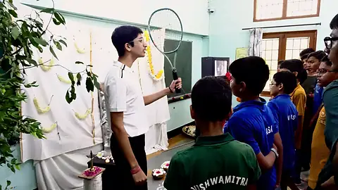 A teacher speaking into a microphone beside a decorated board as students in house shirts look on