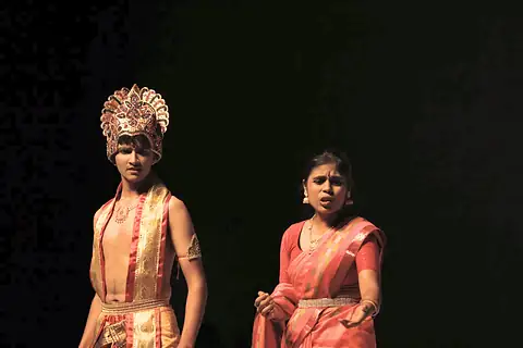 A student in a golden crown and a student in a red sari on a dark stage