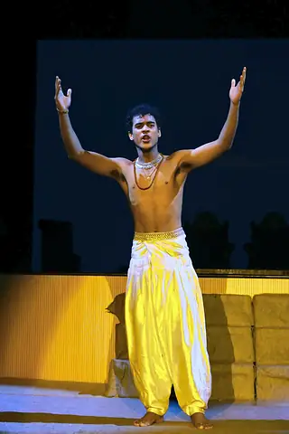 A student in a yellow dhoti alone in a pool of light on a dark stage, both arms raised