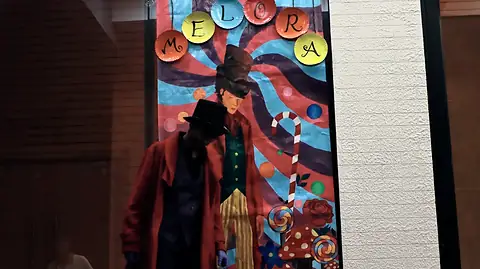 A figure in a red coat and top hat before a painted poster reading Melora