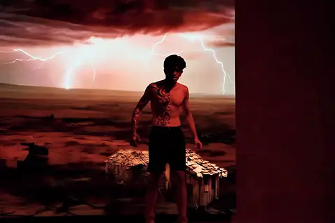 An actor in body paint standing before a projected lightning storm and a broken hut