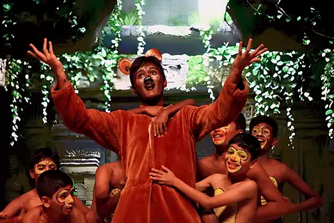 An actor in a bear costume, arms raised, among painted jungle children before a ruined temple set