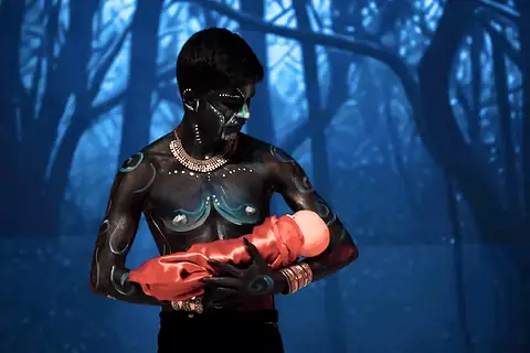 A figure in dark body paint cradling a bundle in red cloth before a blue forest