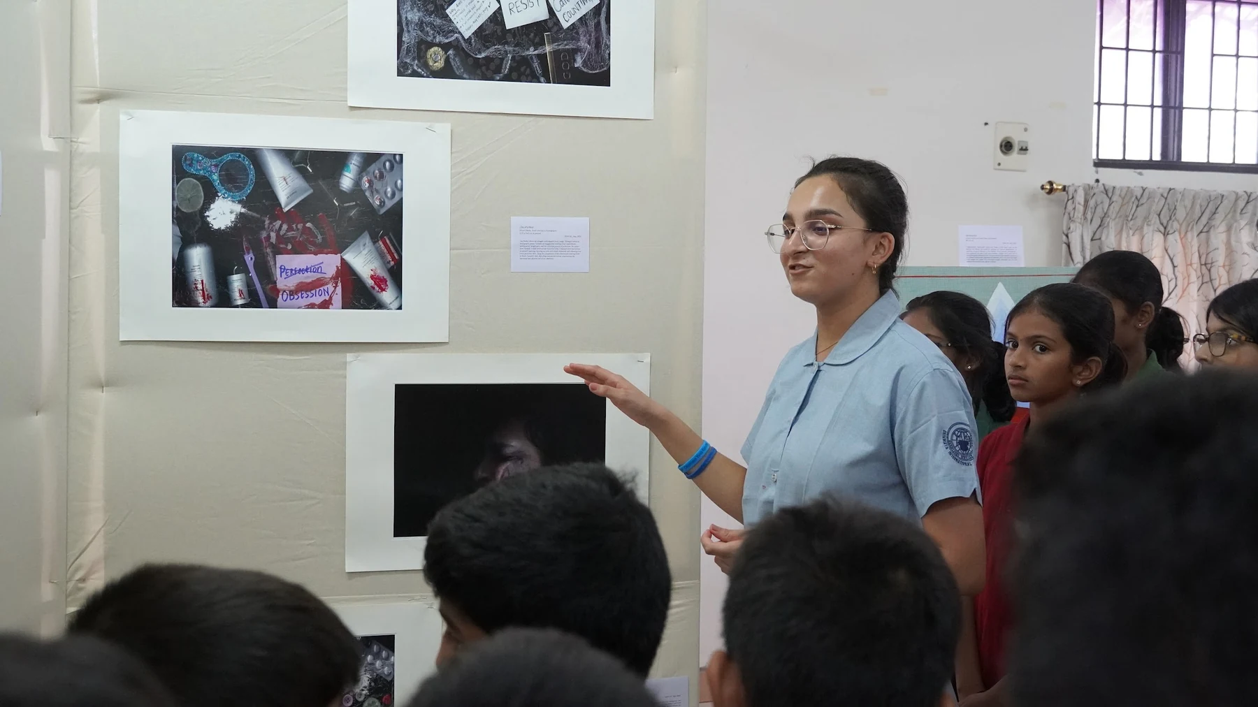 A student gestures towards framed photographs on a display wall as younger students listen