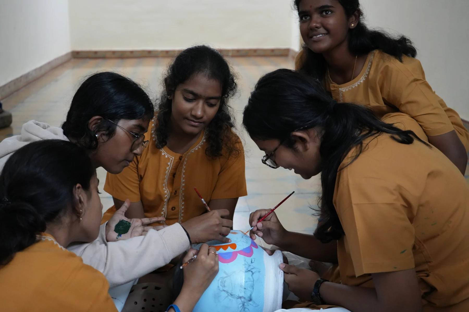 Four students bent over a pale blue clay pot, painting a band of pink and orange with fine brushes