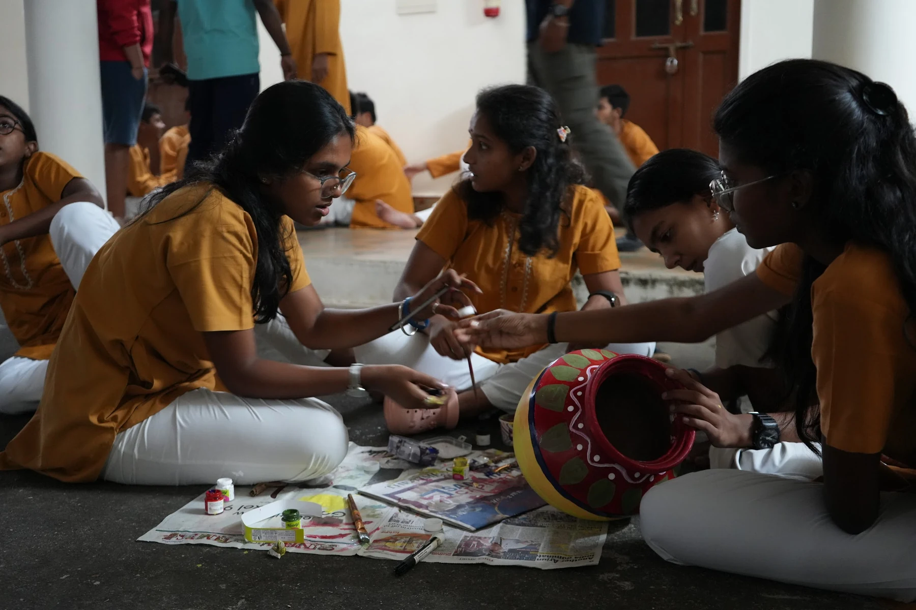 Four students seated on newspaper around a red clay pot painted with green leaves and white dots, one holding a small pink pot and a brush