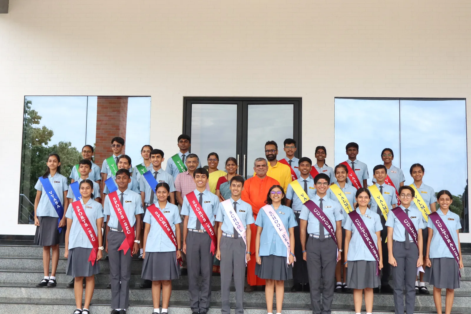 The Student Council 2026–27 standing in rows on the steps of a school building, in sashes over school uniform, with a swami in saffron among them
