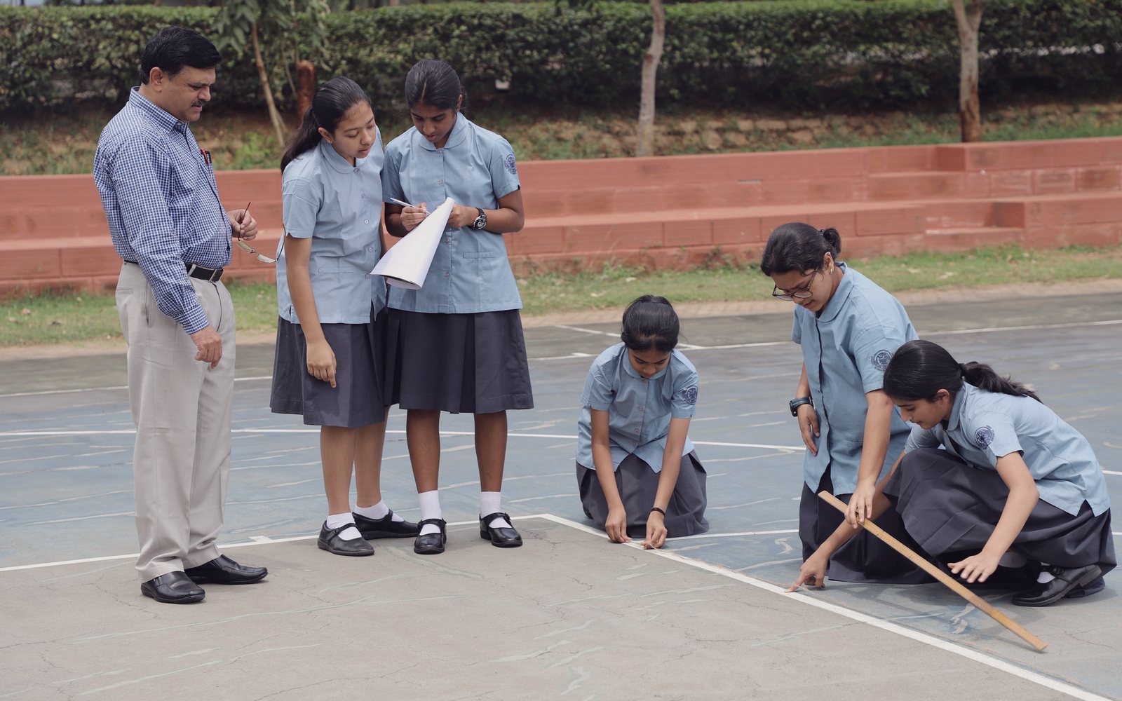 Students measuring out lines on an outdoor court with a long ruler while a teacher looks on