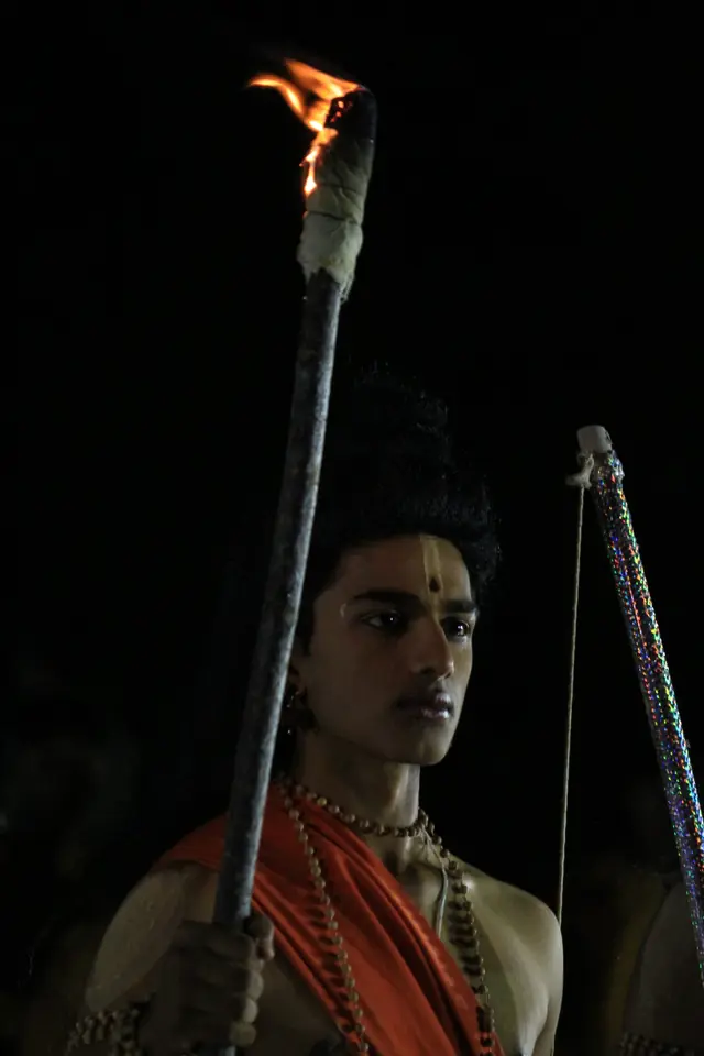 A student in costume as Rama holding a bow beside a burning torch