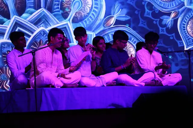A group of students seated on stage singing before a blue projected mandala
