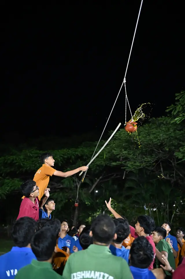 A student on the shoulders of others reaching up for a clay pot hung from a rope, after dark
