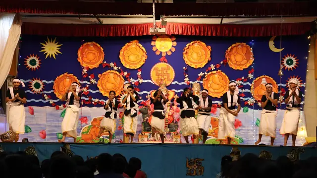 Students in white mundu and dark tops dancing on a stage hung with painted suns