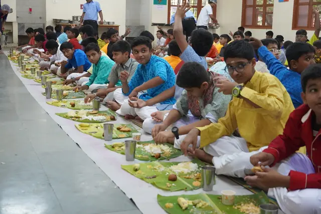 A long row of students seated on the floor eating the sadhya from banana leaves