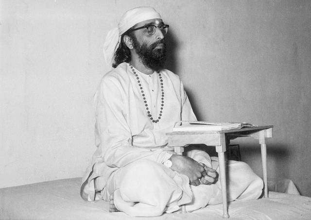 Swami Chinmayananda seated beside a small reading desk.