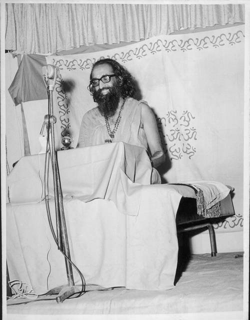 Swami Chinmayananda speaking at a lectern with a microphone.