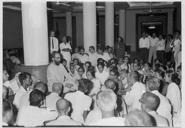 People gathered around Swami Chinmayananda in a columned room.