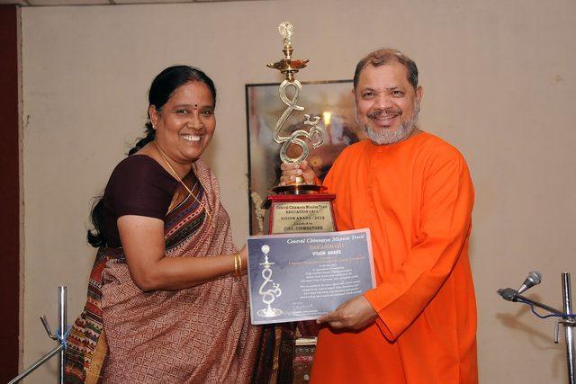 A trophy inscribed 'Central Chinmaya Mission Trust Education Cell Vision Award 2012, awarded to CIRS, Coimbatore', held up with its certificate by a woman in a sari and a swami in saffron