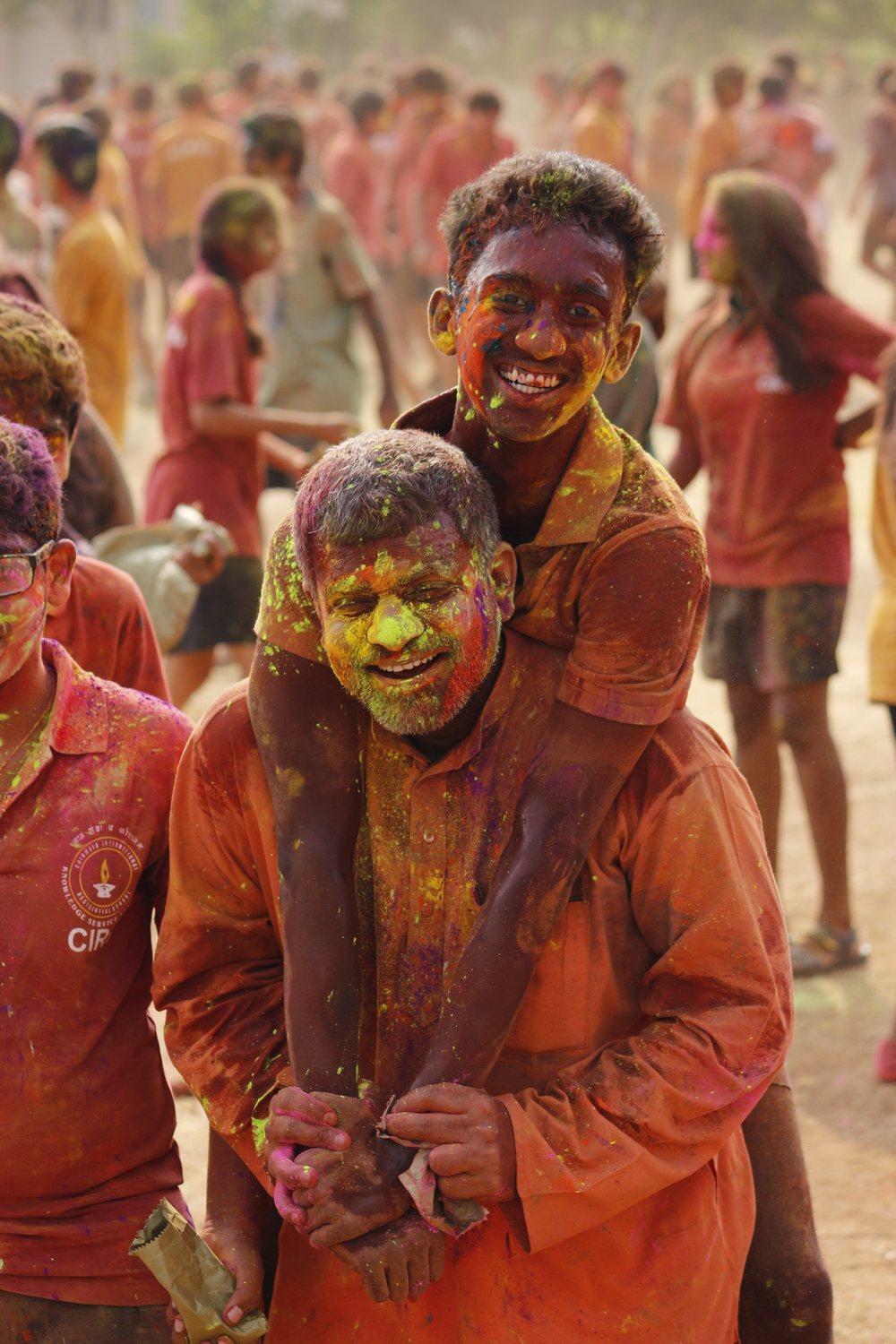 Two students covered in coloured powder, grinning, one riding on the other&rsquo;s back at Holi.