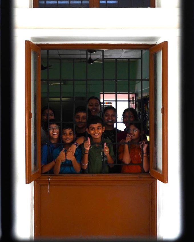 Students crowded behind the bars of an open classroom window, smiling and giving a thumbs up.