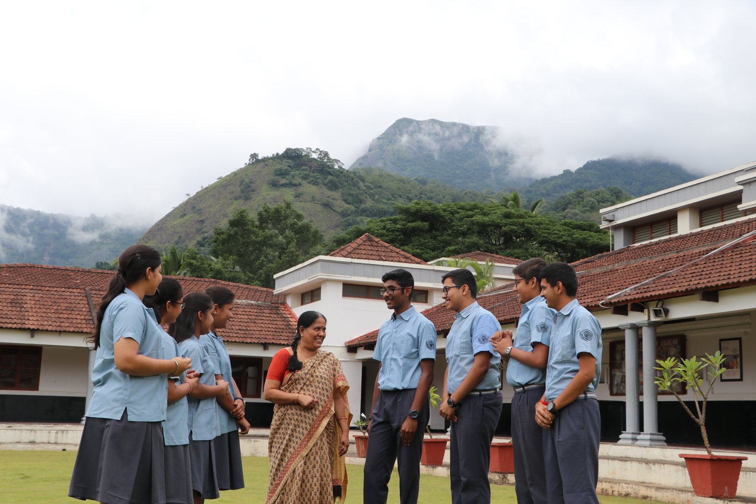 Students in uniform talking with a teacher in a sari on the lawn, tiled roofs and a misty hill behind them.