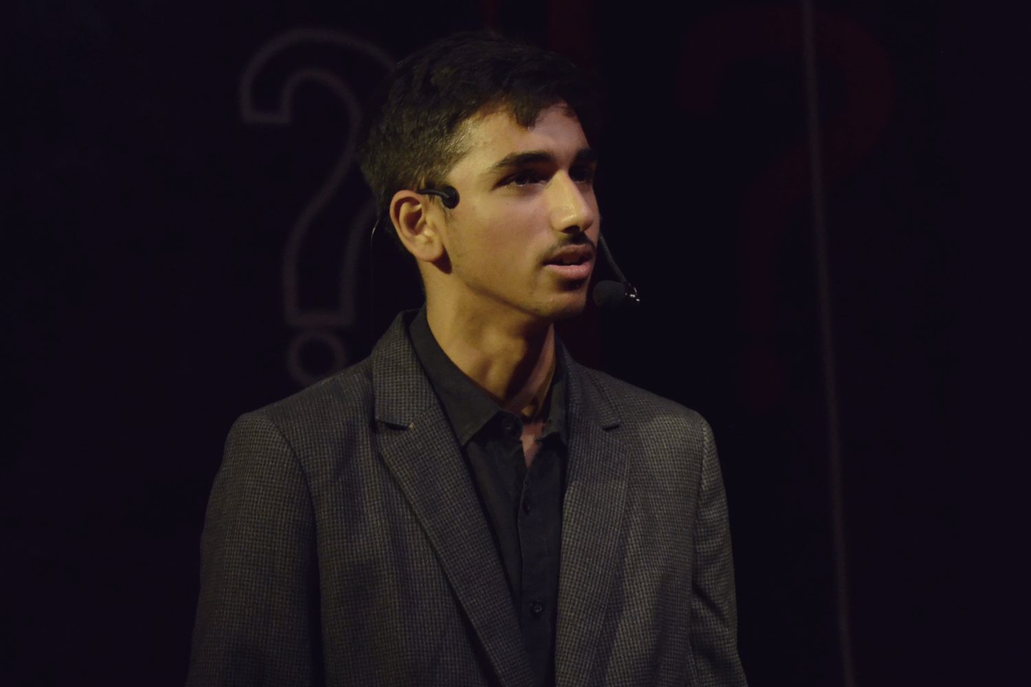 A student on a darkened stage in a headset microphone, a painted question mark behind him.