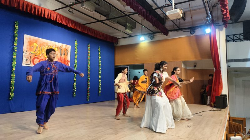 A boy in an indigo costume in mid-step on stage, a group of dancers in white and red behind him