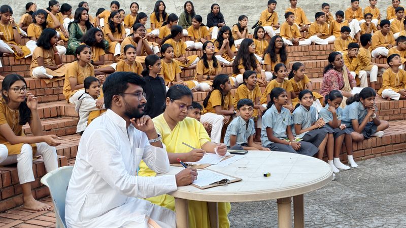Two judges seated at a round table making notes, pupils in mustard kurtas on tiered steps behind them
