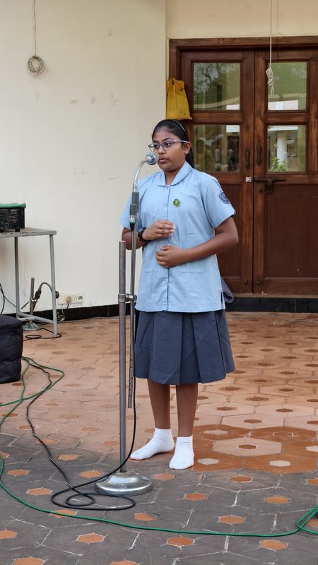 A girl in school uniform reciting at a microphone stand in front of a wooden door