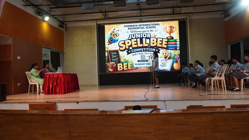 A junior student spelling into a microphone on the stage of the school hall, in front of a screen reading Junior Spell Bee Competition, with two judges at a red-clothed table and pupils seated on the right