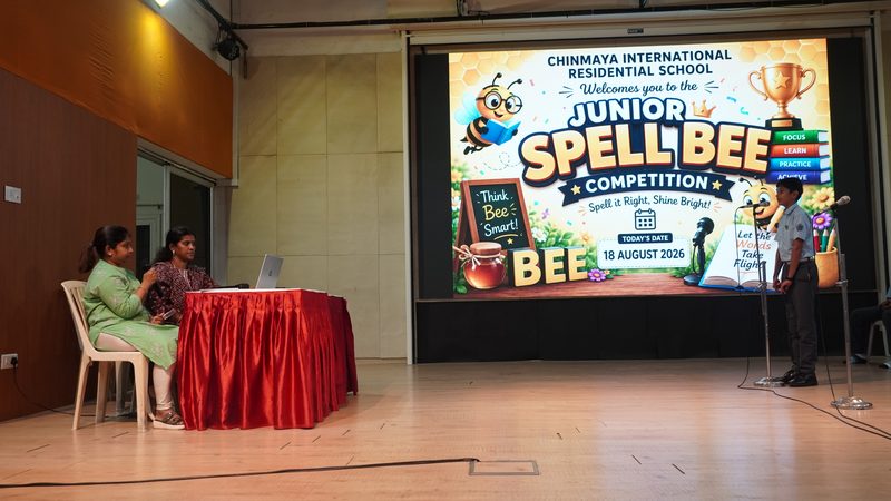 A student at the microphone on stage beside the Junior Spell Bee Competition screen, the judges at their red table to the left