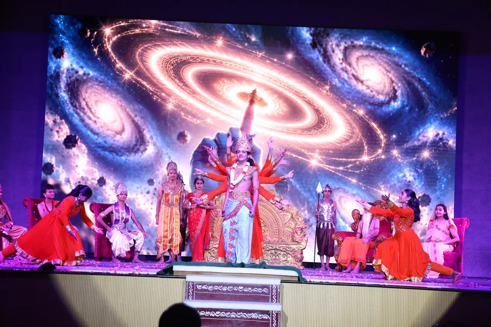 Performers in red and orange costumes on stage, in front of a screen showing a spiral galaxy, with a many-armed figure at the centre