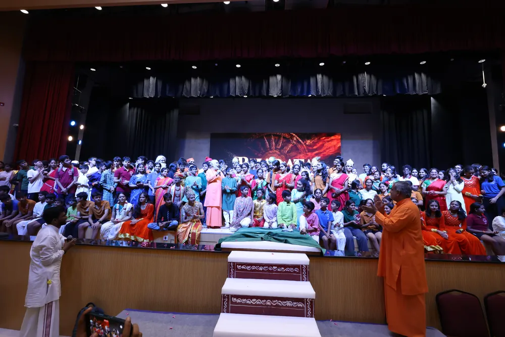 Students in colourful clothes filling the stage, with a swami in saffron standing at the front of it, hands together, and an audience in the foreground