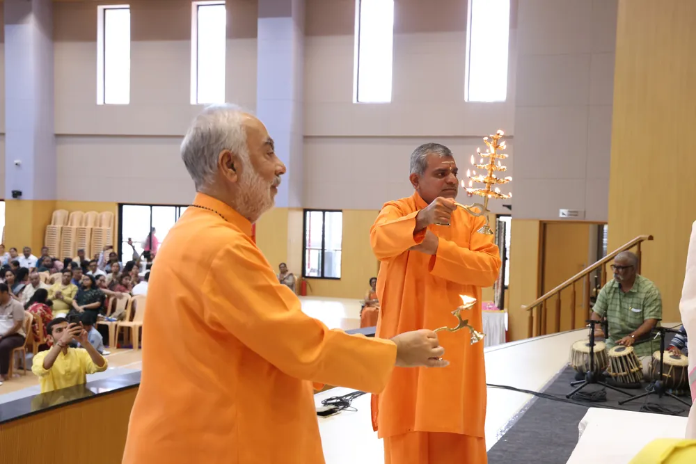 Two swamis in saffron holding brass lamps in front of a seated audience in a hall, with a tabla player beside them
