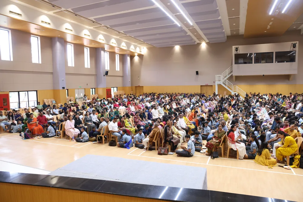 A hall packed with people seated on the floor and on chairs, seen from the front under rows of ceiling lights