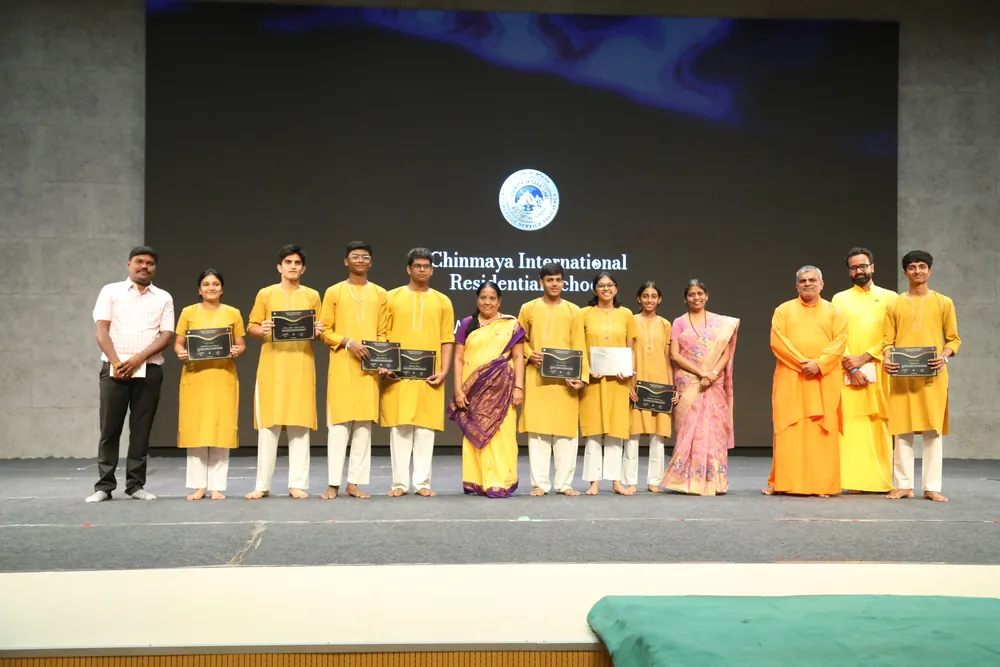 A row of students in yellow kurtas holding dark folders, standing on a stage with teachers and swamis, in front of a screen with the school’s emblem and name