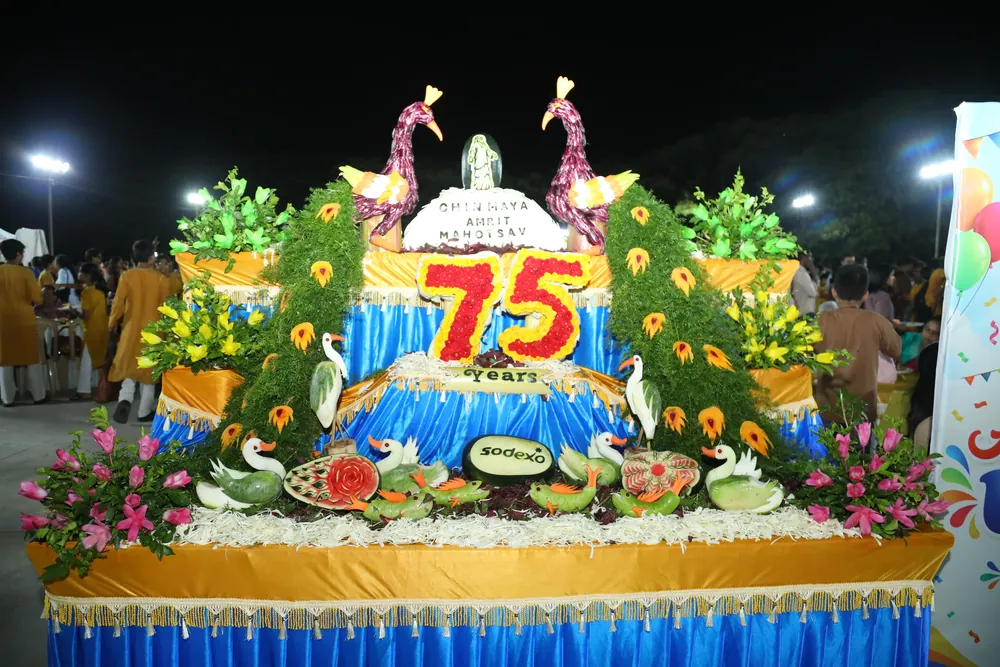 A tiered display of carved fruit and flowers lit at night, with two peacocks at its sides, swans, and the words Chinmaya Amrit Mahotsav and 75 Years