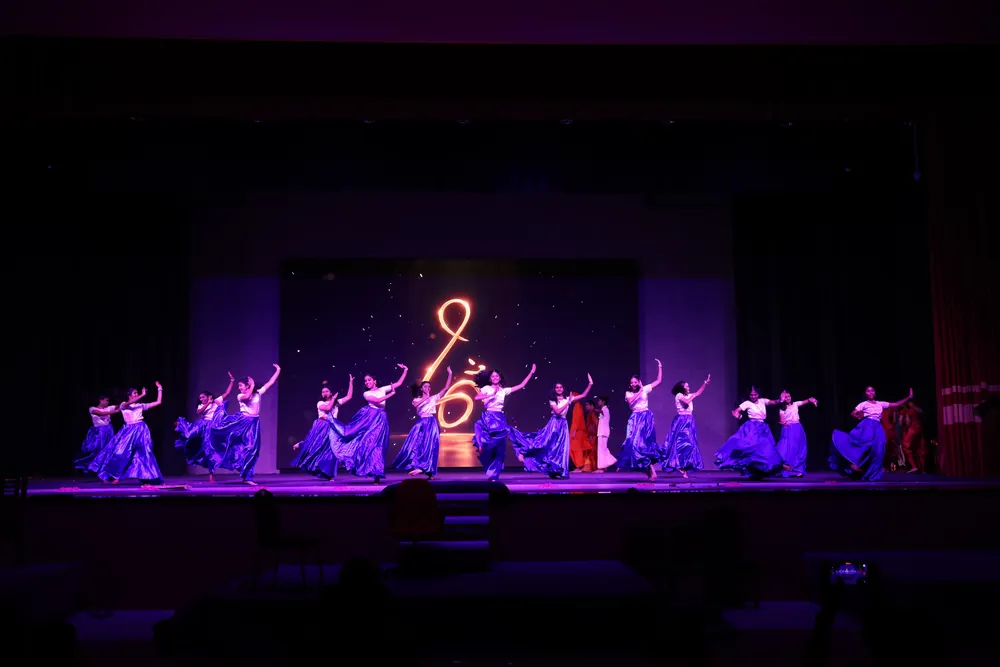 Dancers in blue and purple costumes on a stage lit in violet, with a glowing symbol at the back