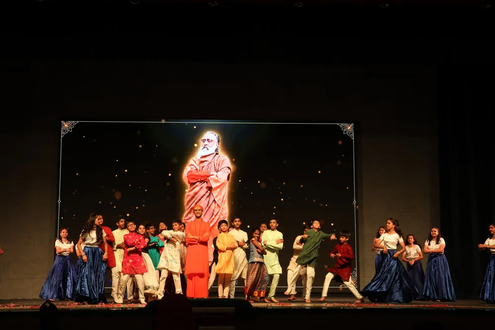 Students in blue skirts and kurtas of many colours on a dark stage, in front of a screen showing a robed figure in saffron