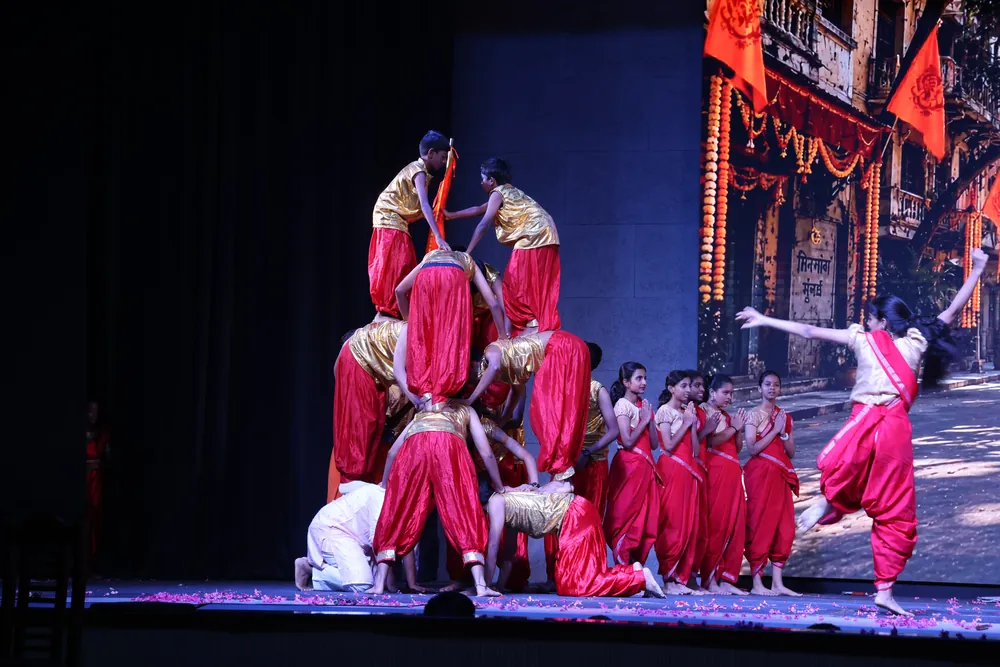 Dancers in red and gold costumes on stage, some of them stacked in a human pyramid, in front of a screen showing a street of garlanded doorways