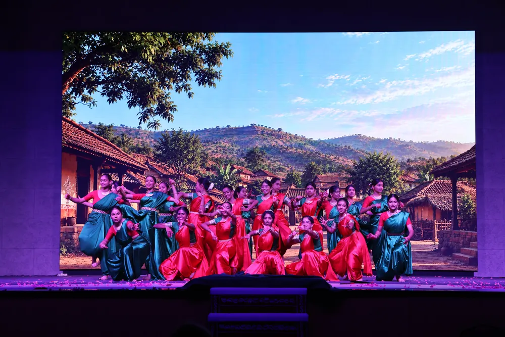 A large group of dancers in red, green and teal costumes posed together on stage, in front of a screen showing a village below green hills