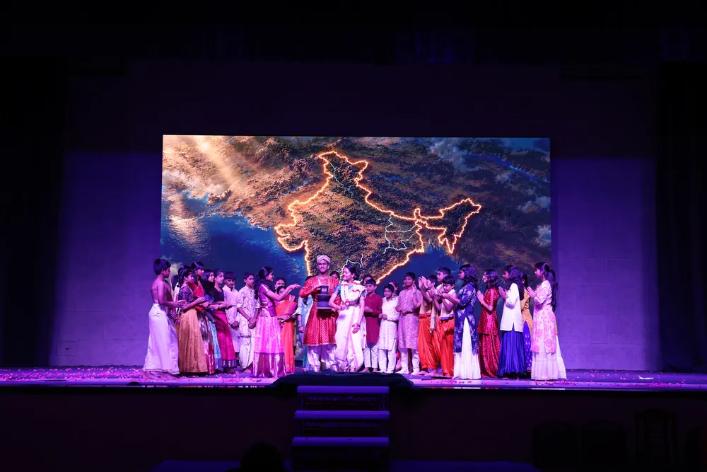 A large group of students in white and coloured clothes on stage, in front of a screen showing a glowing outline map of India