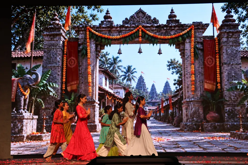Dancers in red, cream and green costumes on stage, in front of a screen showing a carved stone gateway hung with marigold garlands
